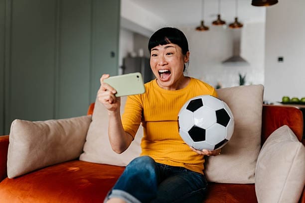 Multiracial woman celebrating a goal and using a smart phone in her living room Woman checking football fixtures on her phone at home